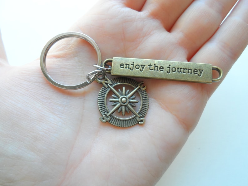 Pode incluir: Um chaveiro de b&uacute;ssola de bronze com as palavras "enjoy the journey" em uma etiqueta retangular. O chaveiro est&aacute; preso a um anel de chave prateado.