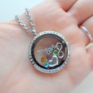 May include: Silver locket necklace with a clear glass insert, featuring a silver infinity symbol, a silver heart with the word "Mimi" inscribed, and colorful crystals. The locket is surrounded by a row of clear crystals.