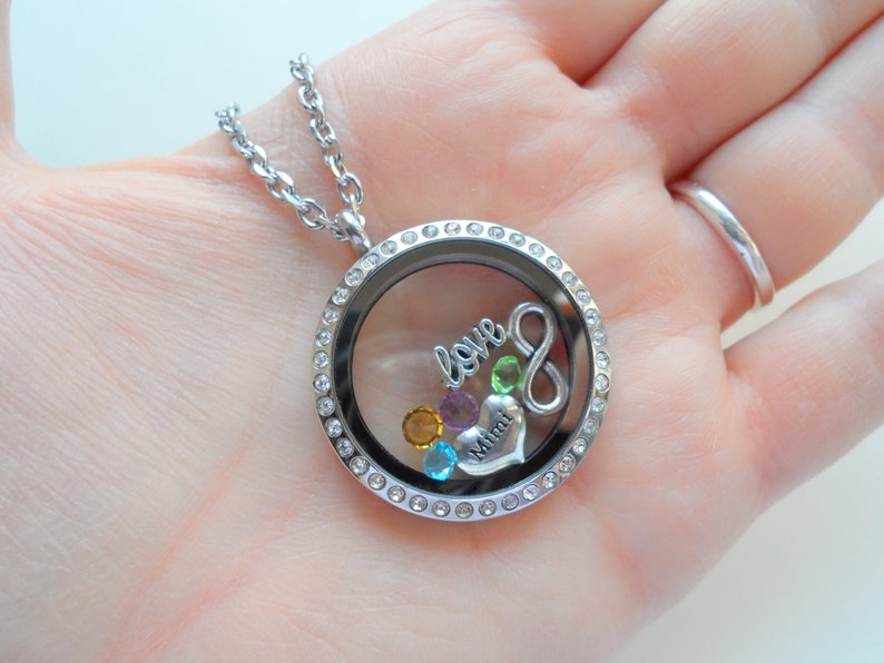 May include: Silver locket necklace with a clear glass insert, featuring a silver infinity symbol, a silver heart with the word "Mimi" inscribed, and colorful crystals. The locket is surrounded by a row of clear crystals.