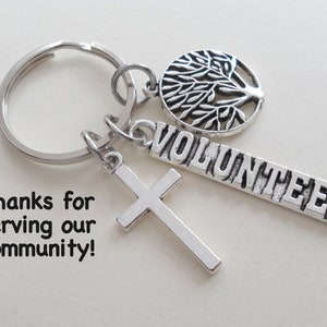 May include: Silver keychain with a cross charm, a tree of life charm, and a rectangular charm that reads "VOLUNTEER". The text "Thanks for serving our community!" is also visible.