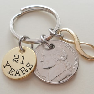 May include: A silver key chain with a gold infinity charm, a gold round charm with the text "21 YEARS" and a silver US quarter coin.