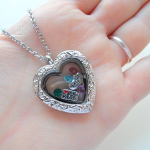 May include: Silver heart-shaped locket necklace with a clear glass window. The locket is filled with small charms, including a red heart with "I love my kids" written on it, a blue charm with "MOM" written on it, and several colorful gemstones.