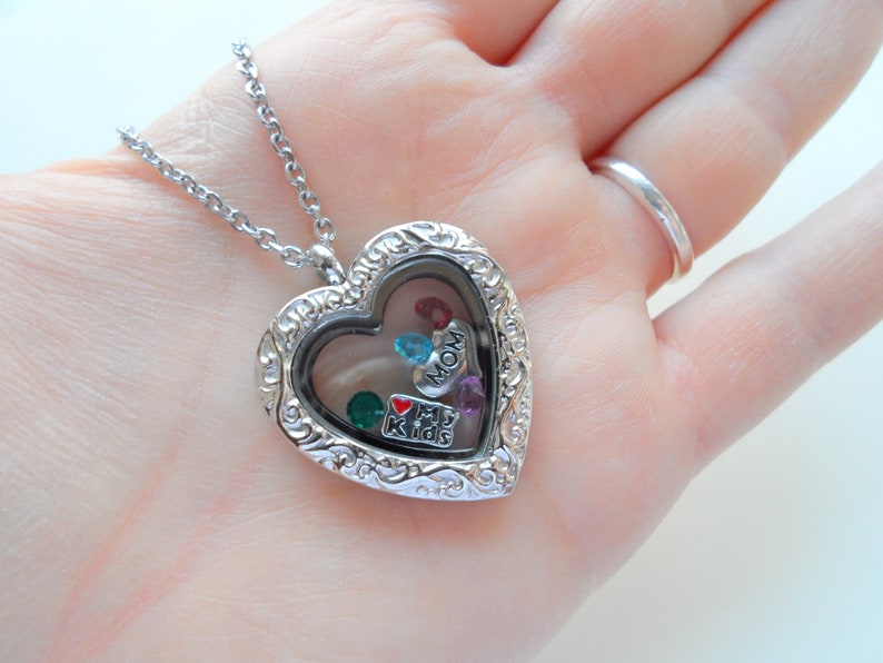 May include: Silver heart-shaped locket necklace with a clear glass window. The locket is filled with small charms, including a red heart with "I love my kids" written on it, a blue charm with "MOM" written on it, and several colorful gemstones.