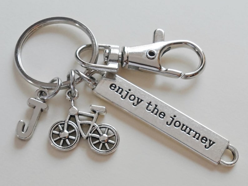 Pu&ograve; includere: Portachiavi color argento con un ciondolo a forma di bicicletta, un ciondolo con la lettera "J" e un'etichetta rettangolare con la scritta "enjoy the journey". Il portachiavi include un moschettone e un portachiavi.