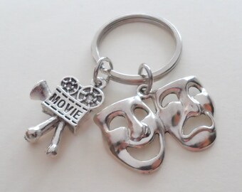 May include: A silver-toned metal keychain featuring two charms attached to a circular ring. One charm is a movie camera with the word MOVIE embossed on its side. The second charm depicts the comedy and tragedy theater masks side by side.