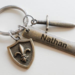 May include: A personalized keychain featuring a shield with a fleur-de-lis, a small sword charm, and a rectangular tag engraved with the name "Nathan". The metal components have a bronze finish and are attached to a silver keyring.