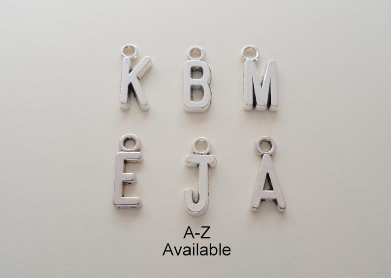 Pu&ograve; includere: Ciondoli in metallo color argento con lettere dell'alfabeto. Sono visibili le lettere K, B, M, E, J e A. Il testo "A-Z Available" &egrave; sotto le lettere. Ogni ciondolo ha un piccolo anello in alto.