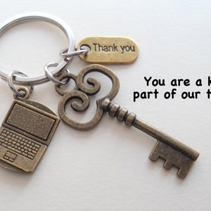 May include: A silver-toned metal keyring featuring three bronze-colored charms: a laptop, an ornate skeleton key, and a rectangular tag engraved with the text Thank you. The text You are a key part of our team! is printed next to the keyring.