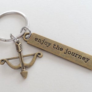 May include: A keychain with a silver ring and a bronze-colored charm. The charm features a bow and arrow design and a rectangular tag with the words "enjoy the journey" engraved on it. The keychain is on a white background.