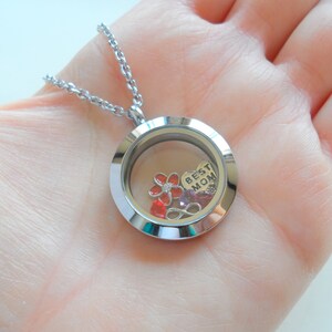 May include: Silver locket necklace with a clear glass insert. Inside the locket are red and pink flowers, a heart charm with the words "Best Mom" and other small charms.