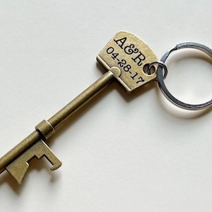 May include: A gold-toned key-shaped bottle opener with a ring attached. The key has a tag with the text "A&R 04-28-17" engraved on it.