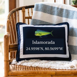 May include: Navy blue decorative pillow with a white border and the word "Islamorada" in white text. A Mahi-Mahi fish illustration is above the text. Coordinates 24.9598°N, 80.5699°W are below the text. The pillow is on a wooden chair.