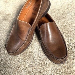 May include: A pair of brown leather loafers with a stitched edge. The loafers have a leather insole with the text "Allen Edmonds" and "Sanibel" printed on it. The text "KZ1 FB 21899 60263" is also printed on the insole.