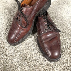 May include: A pair of brown leather dress shoes with black laces. The shoes have a classic design with visible stitching and a dark sole. The leather shows signs of wear, suggesting they are well-used. Other shoes are visible in the background.
