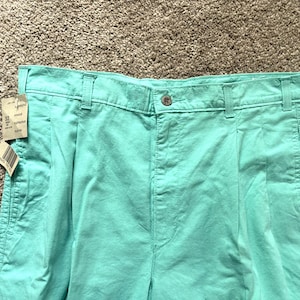 May include: Close-up of turquoise pleated trousers. The trousers have a button closure and belt loops. A tag is attached. The trousers are made of a lightweight fabric, suitable for warm weather. The waist size is 32 inches.