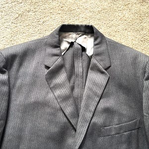 May include: A gray blazer with a subtle striped pattern. The jacket features a notched lapel, a chest pocket, and a two-button closure. The lining is a light gray color.