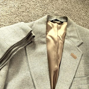 Könnte beinhalten: Ein hellgrauer Blazer mit einem dezenten Karomuster. Die Jacke hat ein goldfarbenes Satinfutter und einen kleinen, rechteckigen Akzent am Revers. Der Ärmel ist umgeschlagen, und der Blazer liegt auf einer strukturierten Oberfläche.