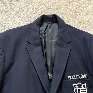Puede incluir: Una chaqueta azul marino con un parche en el bolsillo que dice "Euclid, Ohio" y presenta un escudo con tres flores de lis.