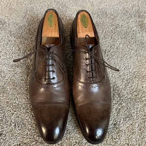 May include: A pair of brown leather Oxford shoes with a cap toe and brogue detailing. The shoes have a lace-up closure and are made of smooth, polished leather. The shoes are on a light brown carpet.