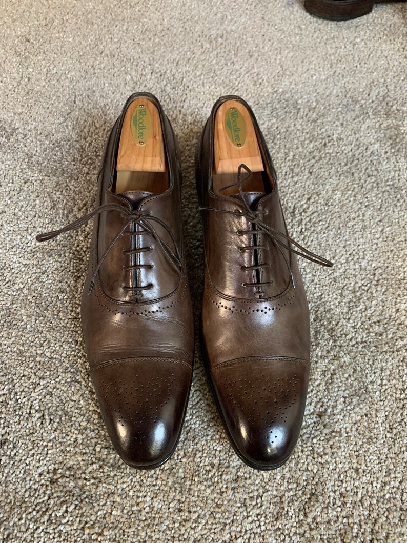 May include: A pair of brown leather Oxford shoes with a cap toe and brogue detailing. The shoes have a lace-up closure and are made of smooth, polished leather. The shoes are on a light brown carpet.