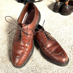 May include: A pair of brown leather dress shoes with laces. The shoes have a classic design with a rounded toe and a stitched sole. Other shoes are visible in the background.