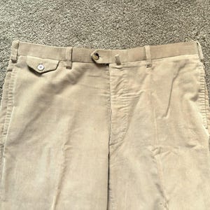 May include: Close-up of beige corduroy trousers. The trousers feature a button closure, belt loops, and pockets. The corduroy fabric has vertical ridges. The trousers are a neutral colour, suitable for various outfits.