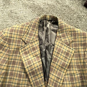 Puede incluir: Una chaqueta de traje de cuadros marrones y amarillos con forro de satén negro. La chaqueta tiene un estilo clásico con solapa de muesca y cierre de dos botones.