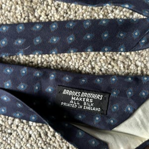 May include: A navy blue silk tie with a white and blue geometric pattern. The tie has a label that reads "Brooks Brothers Makers All Silk Printed in England."