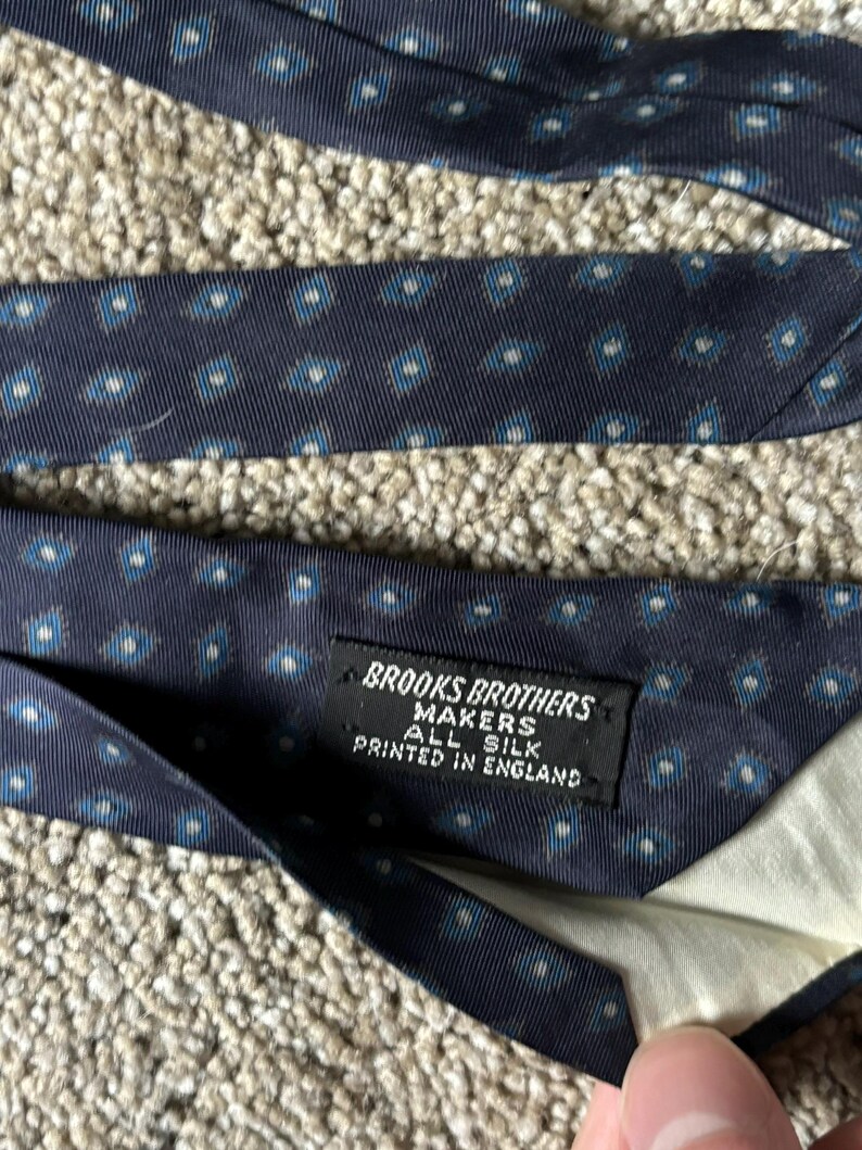 May include: A navy blue silk tie with a white and blue geometric pattern. The tie has a label that reads "Brooks Brothers Makers All Silk Printed in England."