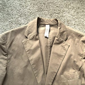 May include: A tan blazer with a notched lapel and a single-button closure. The jacket has a chest pocket and a tag attached to the collar. The fabric appears to be a lightweight cotton or linen blend. The blazer is a neutral color.