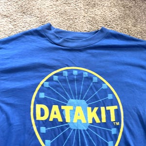 Puede incluir: Una camiseta azul con un diseño gráfico amarillo y azul. El gráfico presenta la palabra "DATAKIT" en amarillo, rodeada de un diseño circular. La camiseta está hecha de un material suave.