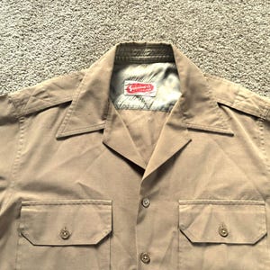 May include: Tan short-sleeve shirt with two buttoned chest pockets and a pointed collar. The shirt has a button-down front and epaulets on the shoulders. A red and white label is visible inside the collar.