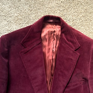 Puede incluir: Primer plano de una blazer de terciopelo burdeos. La blazer tiene solapas con muescas, cierre de un solo botón y un bolsillo en el lado izquierdo. El forro interior es de un tono rojo más claro. La blazer es de estilo clásico.