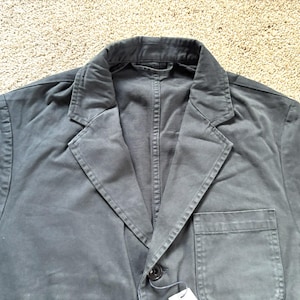 May include: A dark gray cotton blazer with a single button closure and a pocket on the left side.
