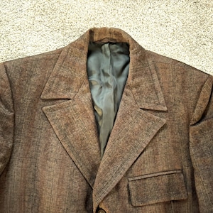 Puede incluir: Una chaqueta marrón con tejido texturizado y un bolsillo en el lado izquierdo. La chaqueta tiene solapa de muesca y cierre de un solo botón.