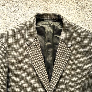 May include: A blazer featuring a small-scale black and white houndstooth pattern. The garment has notched lapels, a structured collar, and a solid black interior lining. The fabric has a textured appearance with a classic tailored construction.