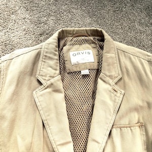 May include: A tan blazer with a notched lapel and a tan mesh lining. The interior features a rectangular label with the text ORVIS and a small graphic. The garment has a single chest pocket on the left side and visible stitching along the lapels.