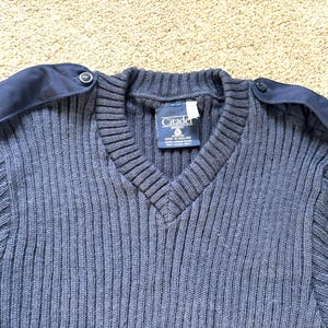 May include: A dark blue knitted sweater with a v-neck and two navy blue shoulder patches with buttons. The sweater has a label that reads "Citadel" and "Made in England".