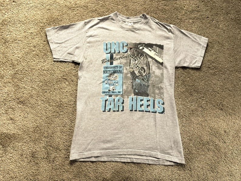 May include: Gray t-shirt with a blue and white graphic print of a basketball player dunking. The graphic includes the text "UNC Basketball, University of North Carolina, Chapel Hill, NC, Tar Heels".