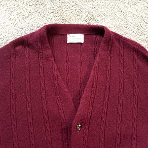 May include: A burgundy cardigan with a V-neck and button fastening. The sweater features a textured, ribbed pattern and a small label near the collar. The material appears to be a soft knit.