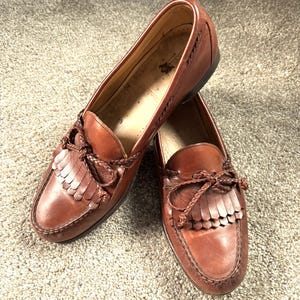 May include: A pair of brown leather tassel loafers with a braided leather strap. The loafers feature a fringe detail and a pointed toe.
