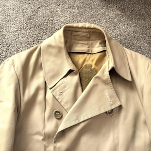 May include: A tan trench coat with a classic double-breasted design. The coat features a notched collar, button closures, and a tailored fit. The fabric appears to be a lightweight material, suitable for transitional weather.