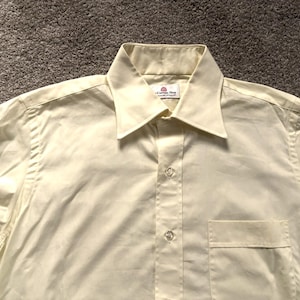 Puede incluir: Camisa abotonada de color amarillo claro con cuello y bolsillo en el pecho. La camisa tiene un diseño clásico con una parte delantera abotonada y un ajuste a medida. La tela parece ser un material liso y tejido.