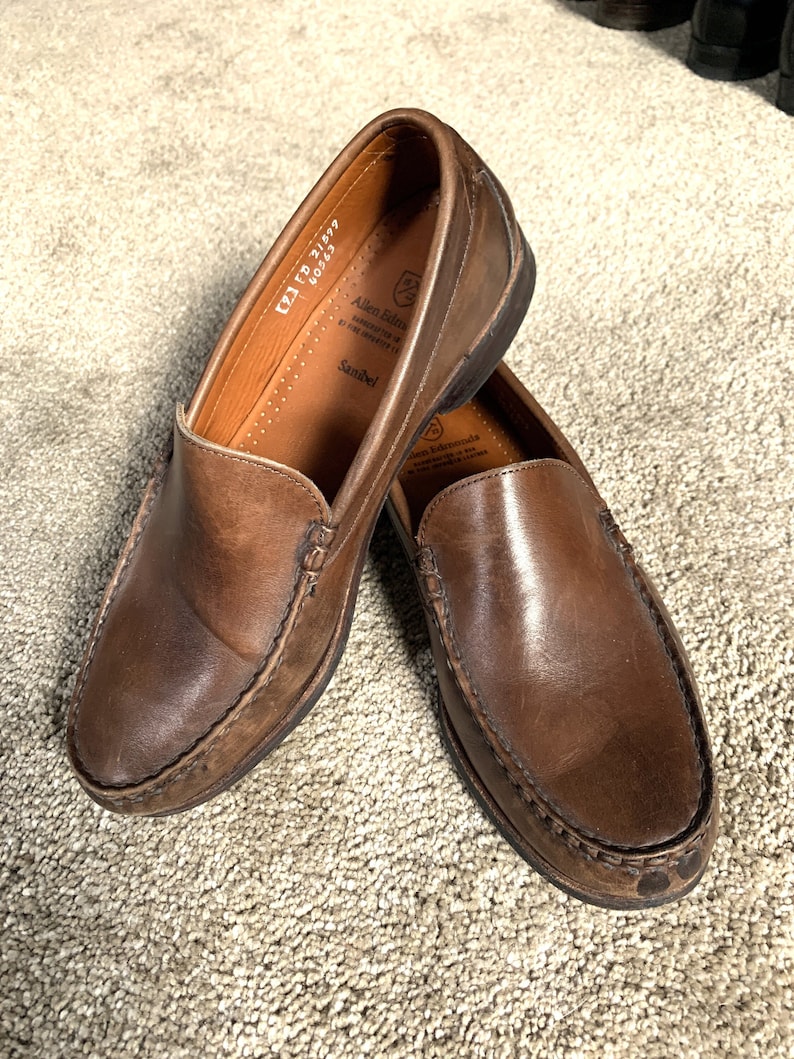 May include: A pair of brown leather loafers with a stitched edge. The loafers have a leather insole with the text "Allen Edmonds" and "Sanibel" printed on it. The text "KZ1 FB 21899 60263" is also printed on the insole.
