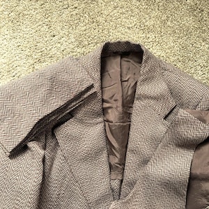 May include: A brown herringbone blazer with a notched lapel and a two-button closure. The blazer has a classic design and a tailored fit. The interior lining is a solid brown color. The blazer is made of a textured fabric.