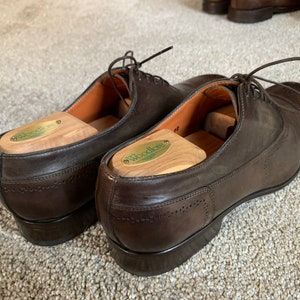 May include: A pair of brown leather dress shoes with shoe trees inside. The shoe trees are made of wood and have the word "Woodlore" printed on them.