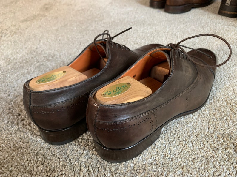 May include: A pair of brown leather dress shoes with shoe trees inside. The shoe trees are made of wood and have the word "Woodlore" printed on them.