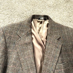 May include: A brown tweed blazer with a subtle plaid pattern. The jacket features a notched lapel, two front pockets, and a light brown satin lining. The fabric has flecks of blue and pink.