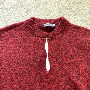 May include: Red and black speckled knit sweater with a single button closure at the neck. The sweater has the brand name "Oakbrook" on the label.