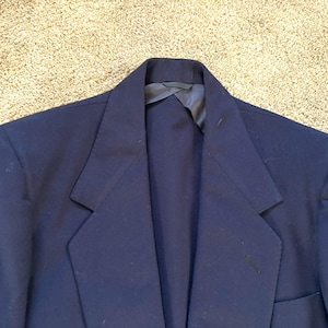 May include: A navy blue blazer with a notched lapel and a single-breasted design. The blazer has a pocket on the right side and a dark lining visible at the collar. The fabric appears to be a smooth, woven material.